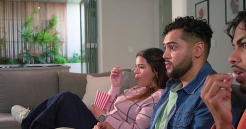 Diverse friends watching movie on cozy living room sofa sharing popcorn, reacting to suspense