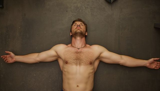 Shirtless muscular man lying supine with arms outstretched on textured workout floor