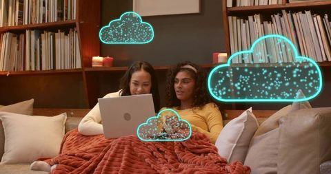 Two women sharing laptop on cozy sofa with neon cloud holograms, candles and blanket