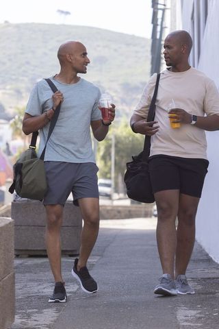 Two african american men walking and talking with gym bags and smoothies on city sidewalk