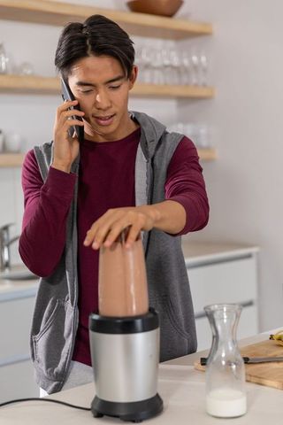 Young asian man talking on phone while making smoothie operating blender in kitchen