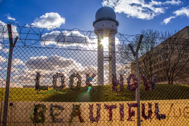 Graffiti is written on a wire fence topped with barbed wire, with an observatory tower and a building in the background. Sunlight is peeking through the structure, creating a bright and dynamic scene. Perfect for themes of urban art, security, creative expression, architecture, or cityscape photography.