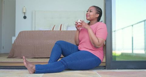 Mid adult african american woman relaxing on doorway holding mug enjoying morning sunlight