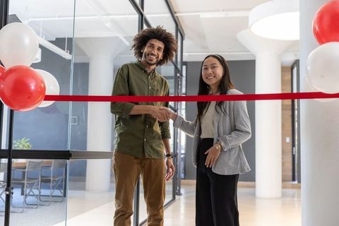 Diverse Coworkers Shaking Hands Across Red Ribbon in Office Lobby