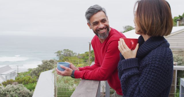 Couple leaning on a wooden railing, enjoying hot drinks and engaging in conversation, overlooking ocean. Ideal for use in lifestyle, travel, vacation, and well-being advertisements, as well as promotional materials for tourism and couple's retreats.