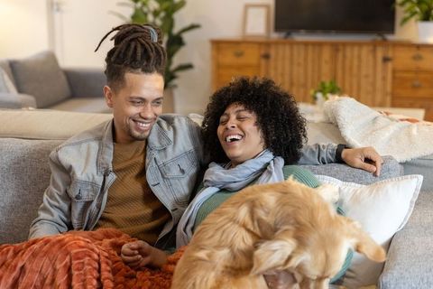 Laughing couple cuddling golden retriever on cozy sectional sofa, relaxing at home