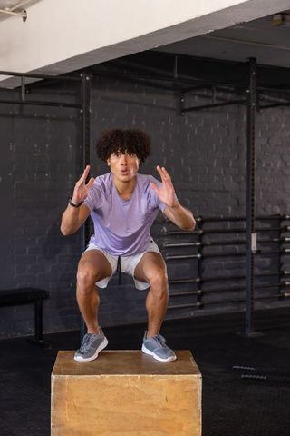 Young athlete performing box squat on wooden plyo box in industrial gym wearing lavender shirt