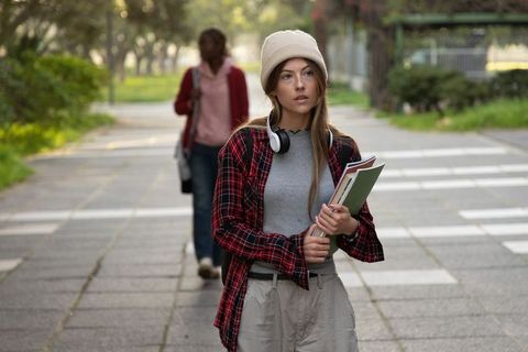 Young white student walking campus path holding notebooks wearing beanie and headphones