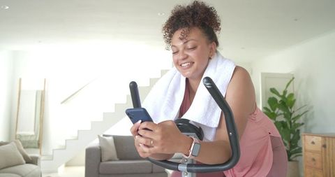 African american woman exercising on stationary bike at home using phone and towel