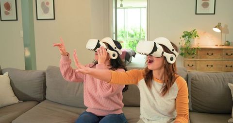 Female friends experiencing immersive VR gaming on sofa reaching forward interacting