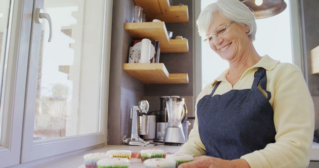 Senior woman wearing glasses and an apron baking cupcakes in a modern kitchen. She is smiling while holding a tray of colorful cupcakes, suggesting a sense of accomplishment and joy in home baking. This image can be used to illustrate themes of retirement, domestic life, culinary hobbies, and the joy of cooking.