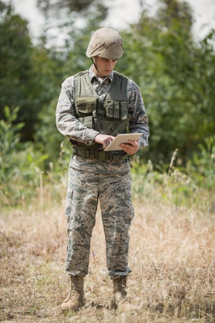 Military soldier in full uniform and tactical gear using a digital tablet outdoors in a boot camp. Ideal for use in articles or advertisements about military technology, modern warfare, training exercises, or communication in the field.