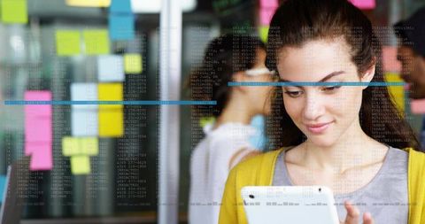 Woman in yellow cardigan using tablet with sticky notes and code on glass wall