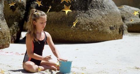 Girl playing with blue bucket on sunny sandy beach near boulders wearing black swimsuit