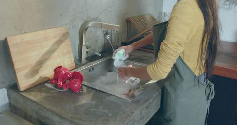 Woman rinsing glass at rustic concrete sink wearing green apron, mustard top, domestic chore