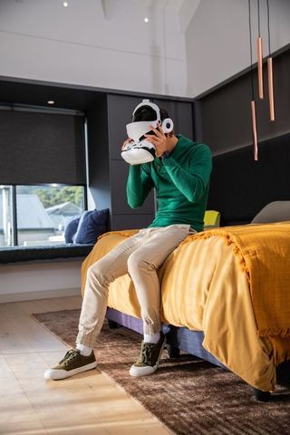 Young man trying virtual reality headset while sitting on bed in modern bedroom