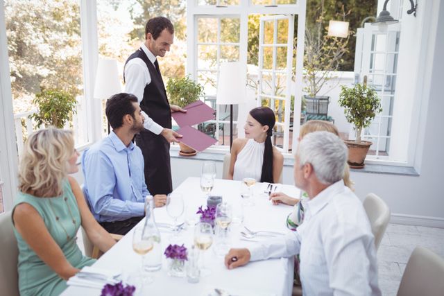 Group of friends sitting at a table in an upscale restaurant, engaging with a waiter while placing their order. Ideal for use in advertisements for dining establishments, social gatherings, or hospitality services. Highlights themes of social interaction, fine dining, and customer service.