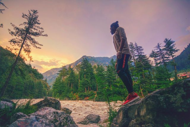 Traveler standing on rocks near a river, admiring scenic view of a forest and mountains at sunset. Ideal for use in travel blogs, adventure promotions, nature conservation campaigns, and outdoor activity advertisements.