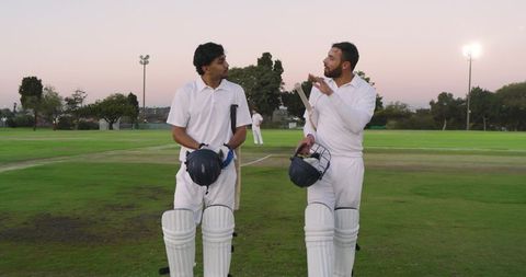 Two cricketers walking and talking at sunset on green cricket field