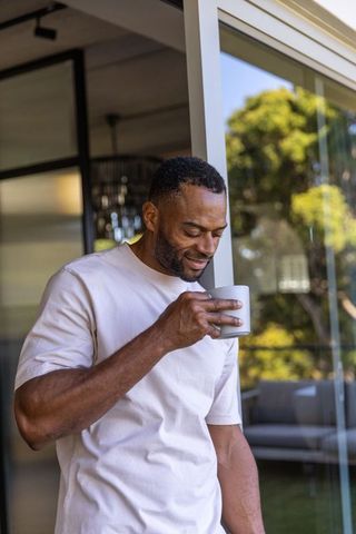 Mature african american man standing at sliding glass door holding mug enjoying sunlight