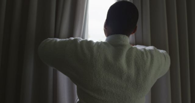 Man in a white bathrobe seen from behind, opening curtains to let in morning light. Ideal for use in wellness, lifestyle, or morning routine themed projects. Suitable for illustrating concepts of relaxation, beginning a new day, or peaceful moments at home.