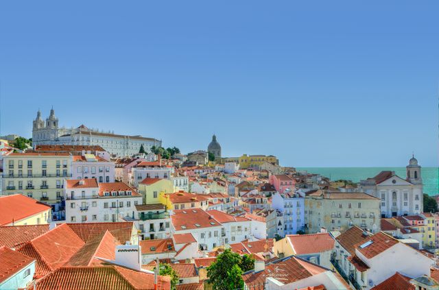 Lisbon’s historic Alfama district viewed from a panoramic angle, featuring rows of charming colorful rooftops beneath a clear blue sky. The scene highlights the architectural beauty and heritage of the city. Ideal for use in travel blogs, websites promoting Portuguese tourism, and print materials related to cultural exploration or Mediterranean destinations.