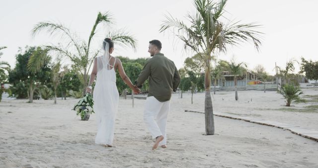 Happy biracial couple walking and holding hands at wedding on beach, copy space. Love, wedding, ceremony, summer and lifestyle, unaltered.