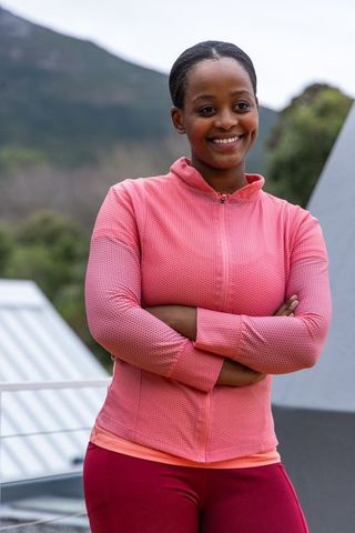 African american woman posing arms crossed smiling in pink workout jacket on rooftop terrace