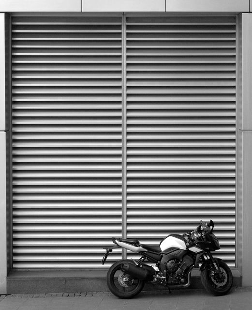 This modern motorcycle stands parked against an industrial metal vent in an urban environment. The sleek design and textured metal vent background create a minimalist and contemporary scene. Ideal for use in automotive, urban lifestyle, or modern design contexts.