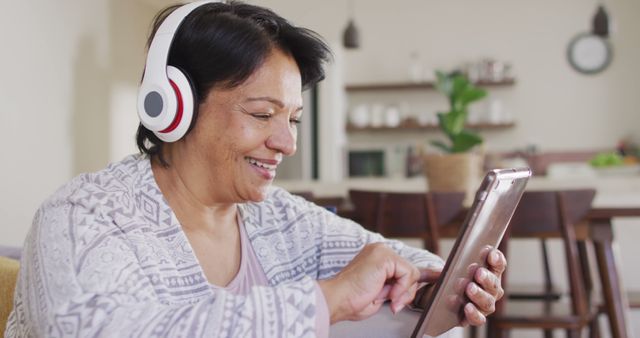 Senior woman is relaxing at home while enjoying music on a tablet wearing headphones. She is smiling and looking content in a casual setting. This can be used in promotional materials for technology, hearing devices, senior living, or lifestyle blogs focusing on senior activities.