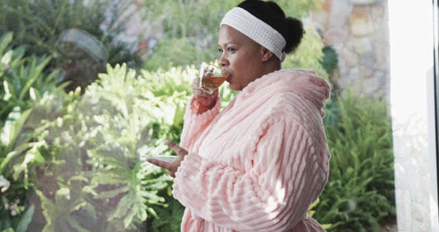 African American woman in pink bathrobe standing near a large window, drinking tea. Perfect for promoting wellness, self-care, and home relaxation. Can be used in lifestyle blogs, morning routine features, relaxation and wellness campaigns.