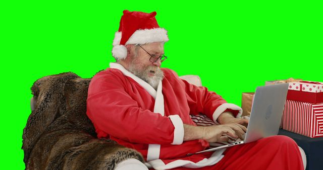 Santa Claus wearing red suit and hat, using a laptop while seated on a couch with a green screen background. Gift boxes are visible around him. Perfect for holiday-themed promotions, virtual background replacements, and festive marketing materials.