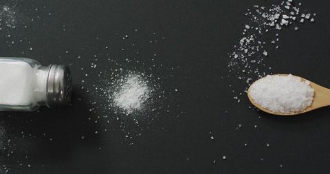 Salt is spilled from a shaker next to a wooden spoon over a dark surface, suggesting a cooking or seasoning theme. Perfect for illustrating concepts related to culinary arts, kitchen essentials, seasoning, or cooking tutorials.