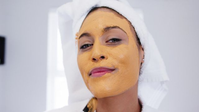 Woman wearing a clay facial mask towel wrapped around her head, smiling contentedly in a modern bathroom. This can be used for beauty and skincare promotions or articles focused on wellness and spa treatments.