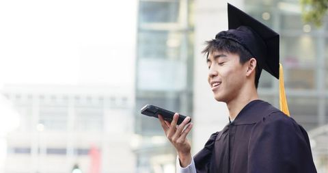 Asian graduate speaking into smartphone on urban campus wearing cap gown with tassel