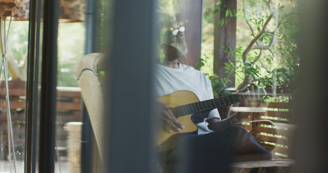 Older man sits comfortably and enjoys playing an acoustic guitar in a serene outdoor setting. Surrounded by nature, he embodies relaxation and tranquility, making it perfect for concepts of leisure activities, hobbies for seniors, and peaceful lifestyle.