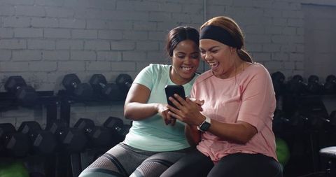 Joyful workout buddies sharing phone and laughter in gym with dumbbells and activewear
