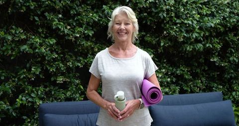 Smiling senior woman holding purple yoga mat and water bottle in green garden