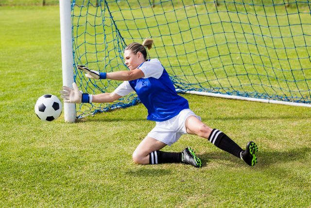 Female goalkeeper in action saving a goal during a soccer game. She is wearing a blue jersey, white shorts, and black socks, diving to her right to stop the ball from entering the net. This image can be used for sports-related content, promoting women's soccer, athletic training materials, or motivational posters.