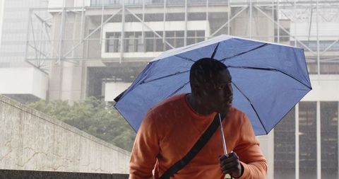 African-American man sheltering under blue umbrella in rainy urban plaza near modern architecture