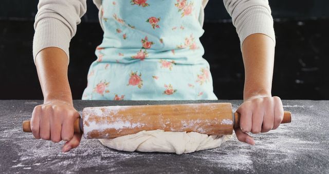 Woman rolling dough on a floured surface in kitchen using wooden rolling pin. Wearing floral-patterned vintage apron, she prepares homemade pastry or bread. Ideal for food blogs, cooking tutorials, and websites focusing on homemade cuisine.