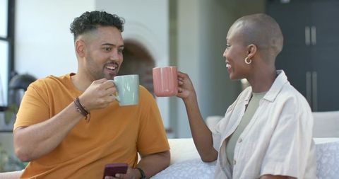 Smiling couple clinking ceramic mugs on cozy sunlit sofa relaxing with smartphone at home