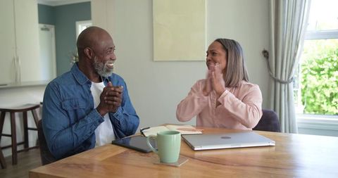 Diverse couple clapping and smiling while celebrating at kitchen table with laptop and tablet