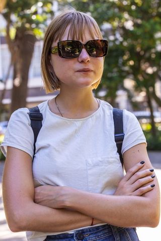 Young woman standing arms crossed wearing sunglasses and casual backpack on sunlit street