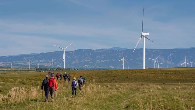 Hiking group trekking toward wind turbines across open grassland with mountain backdrop
