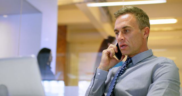 Businessman conversing on phone while working on laptop in modern office environment used for business communication, corporate presentations, and professional working settings.