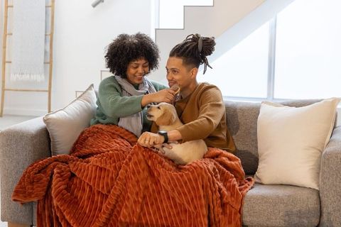 African american couple cuddling with puppy under cozy rust throw on gray sofa