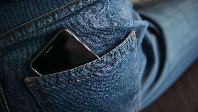 Black smartphone peeking from back pocket of blue denim jeans with orange stitching
