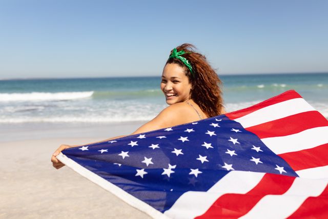 Biracial woman holding American flag at beach, looking back. She has curly brown hair, light brown skin, and is wearing green headband, unaltered