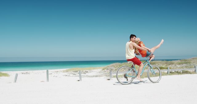 Couple enjoying a playful moment riding a bicycle together on a sunny beach, against a backdrop of clear blue skies and turquoise ocean. Perfect for use in travel brochures, summer vacation promotions, advertisements targeting youth or romantic getaways, and lifestyle magazines.